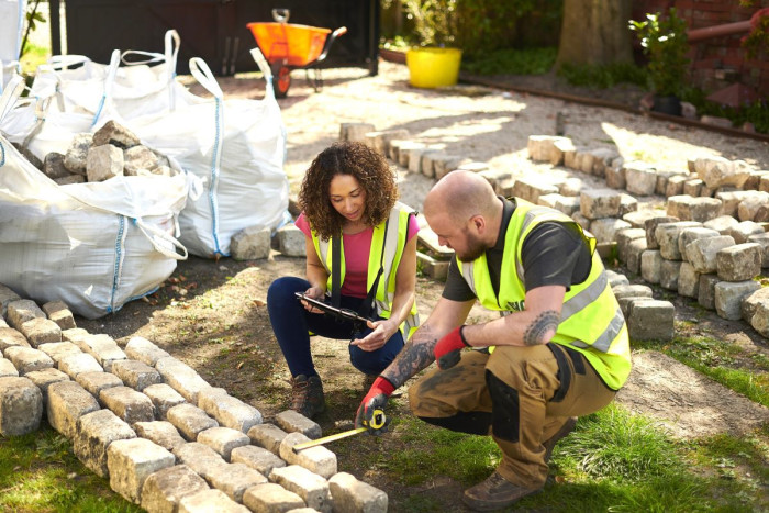 Two landscapers squat down on a lawn surrounded by paving stones
