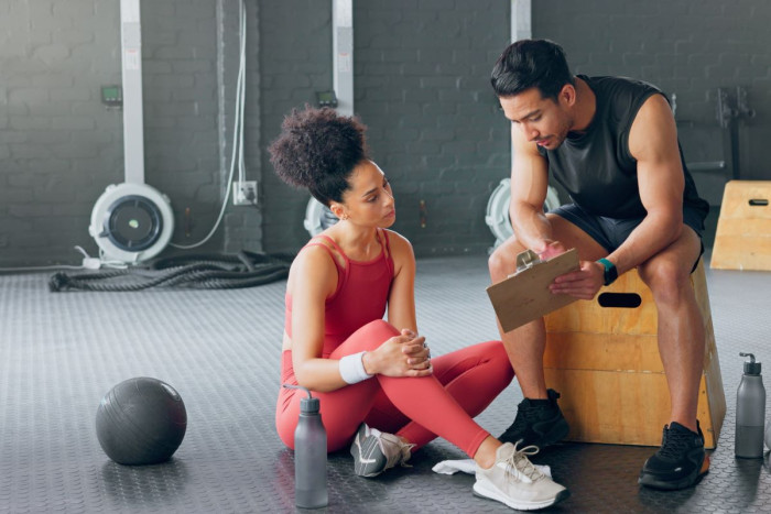 A young woman and a young man in gym clothes sit in a gym, he is shoing her a folder 