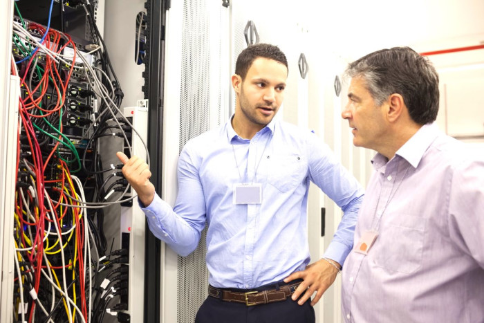 Two men in business clothes stand talking outside a server room full of wires